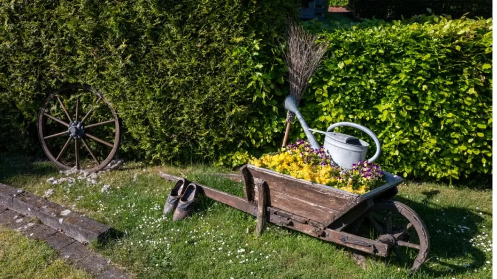 Jardim sutentável frente a uma cerca vica, carrinhola com flores e um regador de metal.