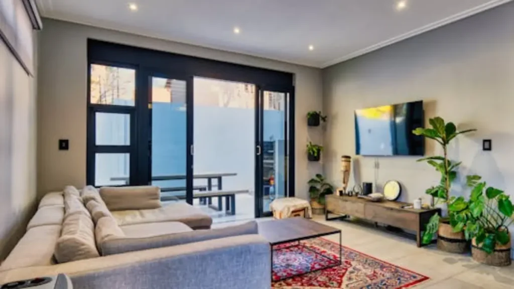 Sala de estar moderna com sofá cinza em L, TV de tela plana na parede, portas de vidro pretas para o exterior, tapete vermelho e plantas verdes.