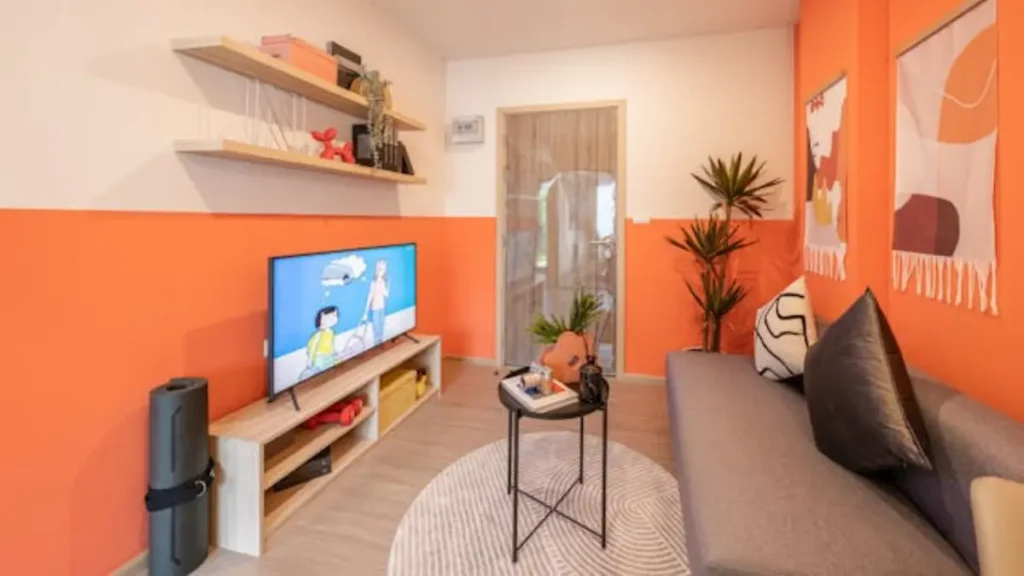 Sala de estar colorida com parede laranja, sofá cinza, TV e prateleiras de madeira.