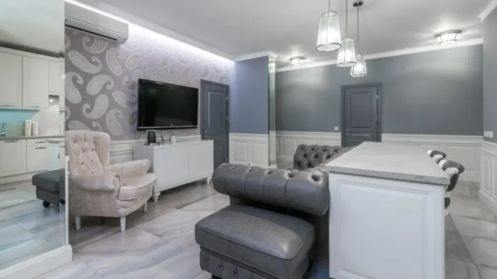 Sala de estar elegante com sofá Chesterfield cinza, poltrona bege, TV na parede com papel de parede e cozinha integrada.