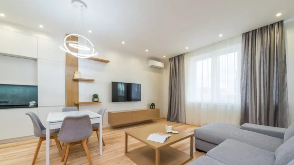 Sala de estar integrada com sofá cinza em L, mesa de jantar branca, TV na parede e cortinas cinza.
