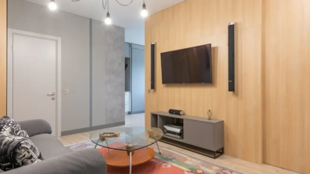 Sala de estar moderna com papel parede imitando cor de madeira, TV, sofá cinza e mesa de centro de vidro.