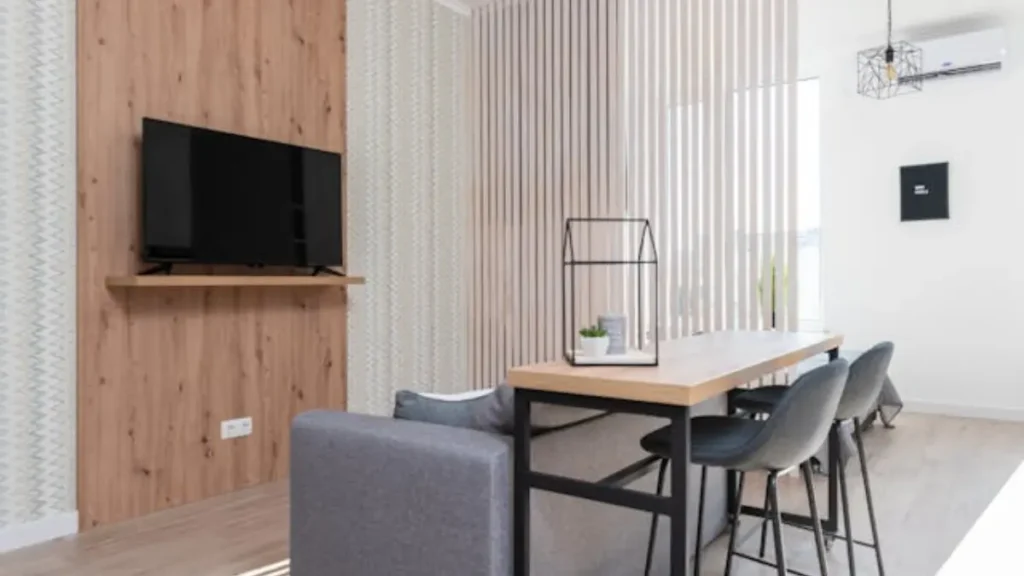 Sala de estar moderna com parede destaque de madeira, TV, sofá cinza e mesa alta com banquetas.