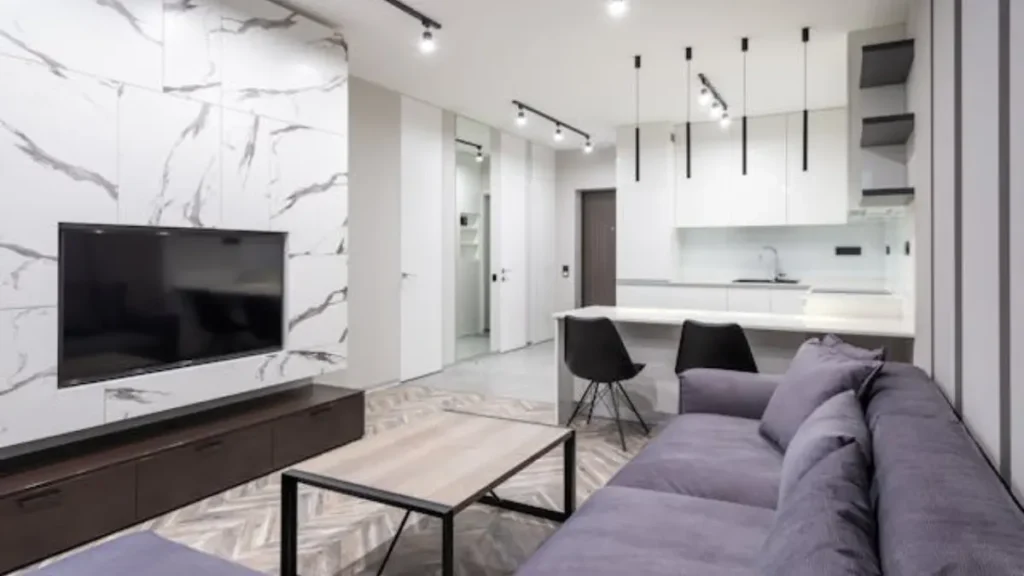 Sala de estar moderna com TV em painel de mármore branco, sofá cinza, mesa de centro e cozinha integrada.