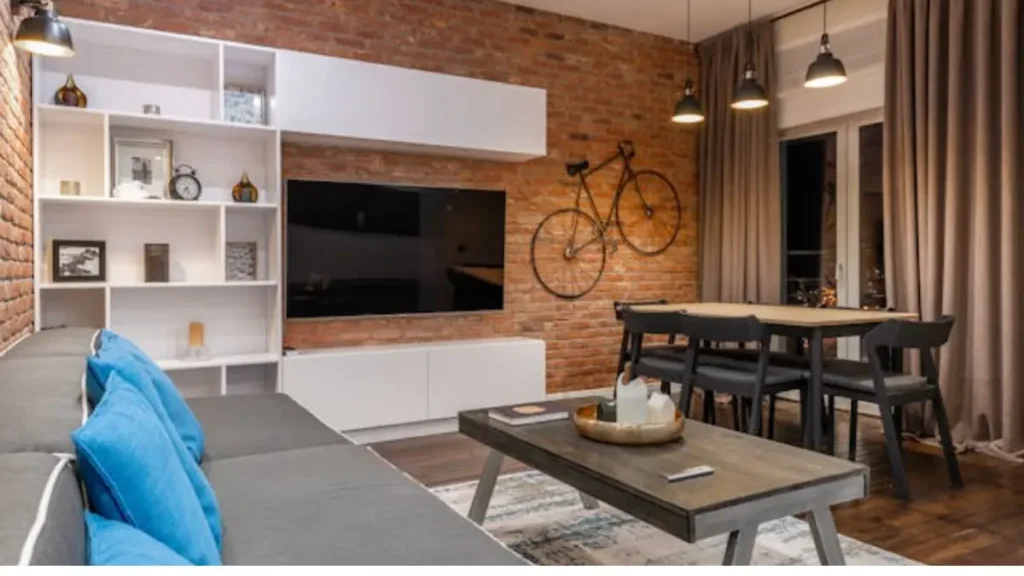 Sala de estar com parede de tijolo aparente, sofá cinza com almofadas azuis, TV, mesa de jantar e bicicleta decorativa na parede.