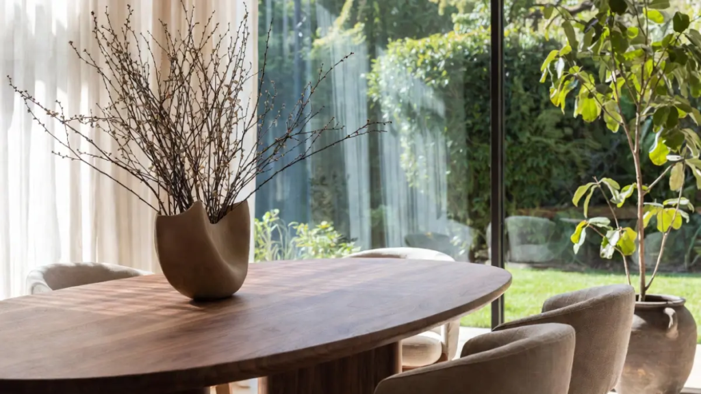 Mesa de jantar oval em nogueira com cadeiras em linho, vaso cerâmico e janela grande com vista para o jardim