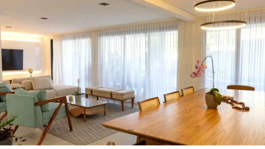 sala moderna com luz solar da manha intensa através de parede toda de vidro com cortinas tranparentes de vool. Mesa de madeira clara, poltronas em tom menta, sofá neutro e luminárias circulares suspensas.